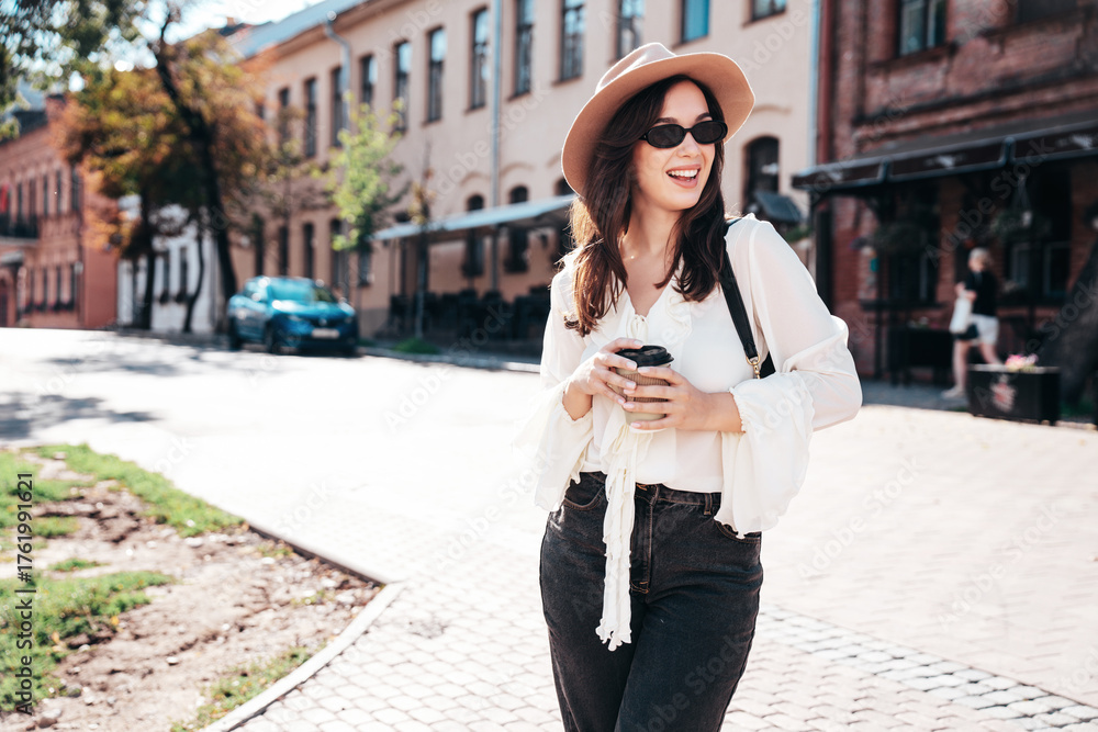 Obraz premium Lifestyle portrait of young stylish woman dressed in white blouse. Model walking with coffee cup in old town in sunny day. Beautiful smiling female in hat. Cheerful and happy. Takeaway coffee
