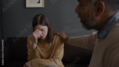 Front view shot of young woman crying during therapy session while sitting on couch with caring therapist supporting client, copy space