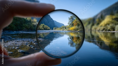 Hand holds a clear lens, framing a vivid river scene between mountains. The lens sharpens the view