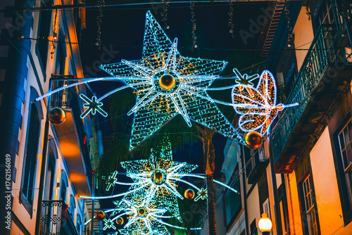 Street at night decorated with festive Christmas lights. Large star-shaped and butterfly-shaped illuminations hang above the street, brightening the dark buildings