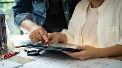 A man and a woman are looking at a tablet with a finger pointing at something. The tablet is on a table with a laptop and a book nearby. Scene is collaborative and focused