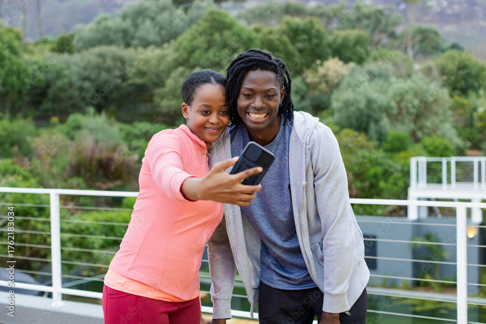 Naklejka premium African American friends taking selfie on terrace overlooking garden in sportswear with smartphone