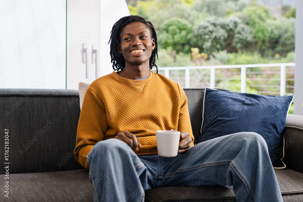 Fototapeta premium African American man sitting in living room on sofa holding mug smiling by sliding glass doors
