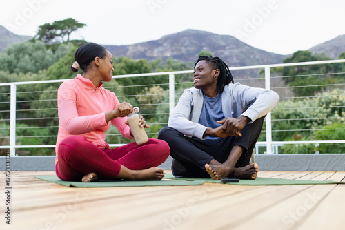 African American friends twisting open reusable water bottle sitting on yoga mats on wooden terrace