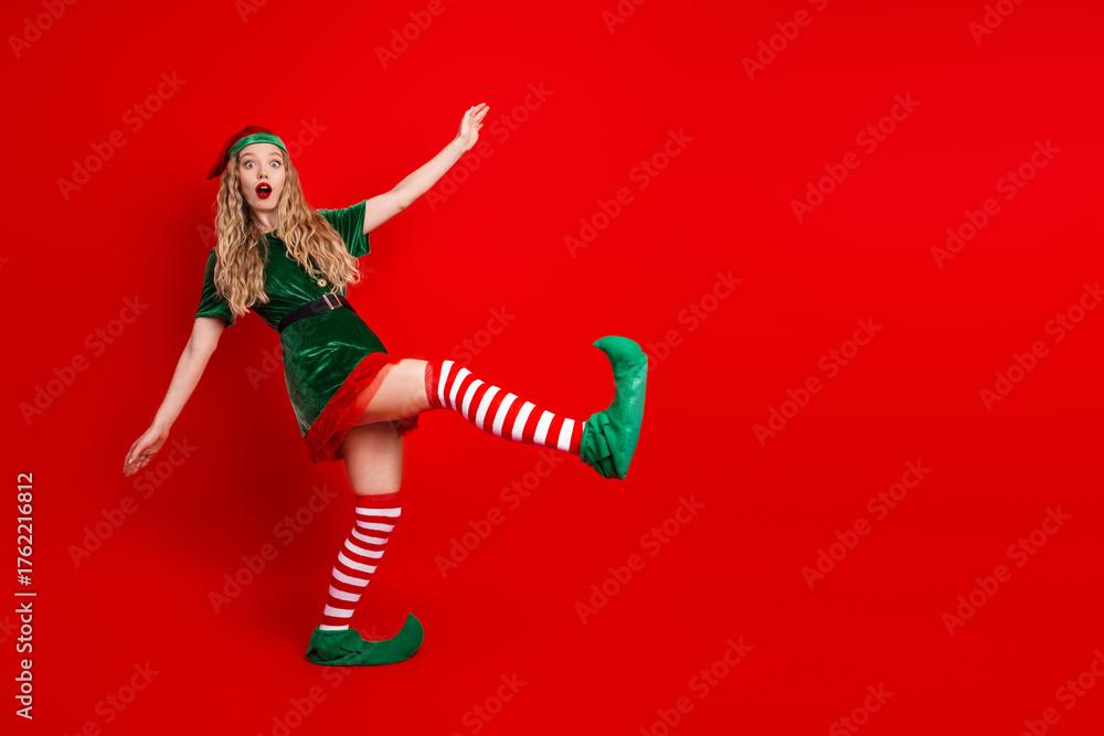 Naklejka premium Festive young woman in a cheerful elf costume striking a playful pose on a vibrant red background for Christmas celebration