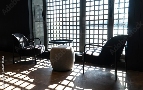 Two soft chairs and a table near a large window with bars that cast shadows on the floor 