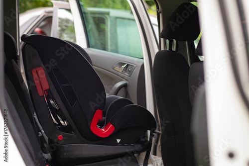 A close-up shot of a black child car seat with bright red latch elements, installed on the rear seat of a white car.