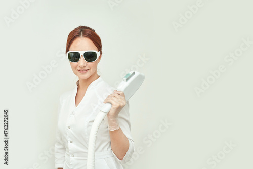 Female caucasian adult holding laser hair removal device in white attire and protective glasses.