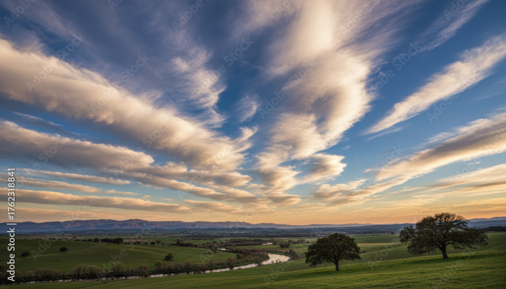 Fototapeta premium Swirling clouds stretch across the sky as the sun sets gently behind distant mountains. A winding river flows through the lush valley, framed by trees on either side
