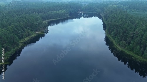Slow forward drone shot over a calm, dark lake surrounded by dense evergreen forest on a misty, overcast morning. Reflection of the sky on water. Kashubia, Pomeranian, Poland