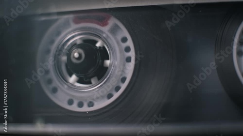 Close-up view of a classic audio cassette tape rewinding in a player. The white plastic reel with circular holes spins, winding the magnetic tape on the spool inside the transparent housing.
