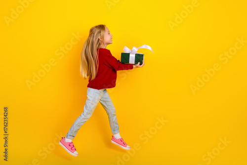 Playful young girl in red sweater carries a green gift box with white ribbon against a bright yellow background in a joyful holiday moment