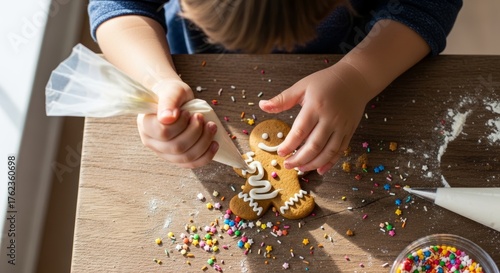 A close-up, overhead shot of a child's hands using a piping bag to decorate a gingerbread man cookie with white icing and colorful sprinkles on a wooden table.