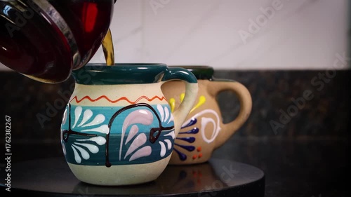 Hot black coffee being poured from a glass pot into a colorful, handmade ceramic mug in a kitchen. mexican coffee