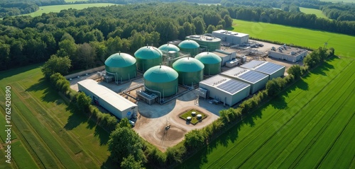 Aerial view of biogas plant with green tanks in countryside surrounded by trees, fields. Renewable energy facility produces electricity from organic waste, agricultural residues. Solar panels on