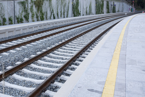 Railway tracks stretch into the distance, bordered by a clean concrete platform, with yellow tactile paving and greenery along the walls, illustrating urban transportation infrastructure