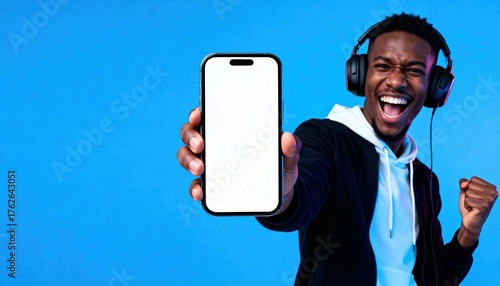 Emotional African American guy showing smartphone. Happy man with headphones wins, holding phone with blank screen for mockup. Mobile app advertisement concept on blue background.