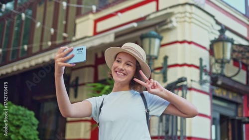 Cute traveler girl in straw hat with backpack on city street taking selfie on phone with tourist hotel background for social media