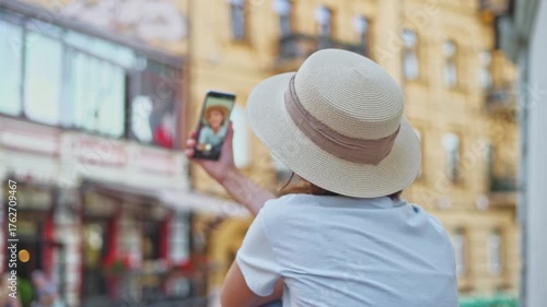 Pretty young blogger tourist girl in straw hat making live stream on phone for social media audience blogging about tourism and travel with tourist city background back view