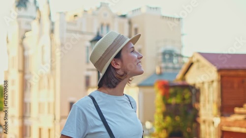 Smiling tourist girl in straw hat on summer city street happily examining tourist city architecture sunny day during travel