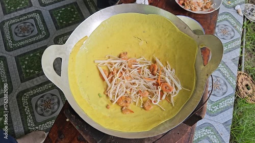 Close-up of a Vietnamese sizzling pancake (banh xeo) with shrimp, bean sprouts, and carrots in a hot pan.	
