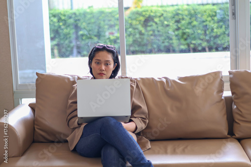 Asian woman sitting on sofa using laptop for online business or working from home.