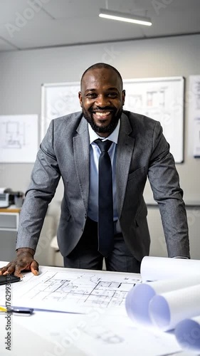 Smiling man in a suit leans on a desk with blueprints in an office, another person working in background