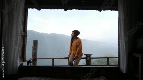 A woman sitting on wooden balcony with mountain views on foggy morning outside the window