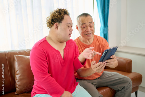 A man with down syndrome and senior man sharing an excited moment together while looking at a tablet on a sofa
