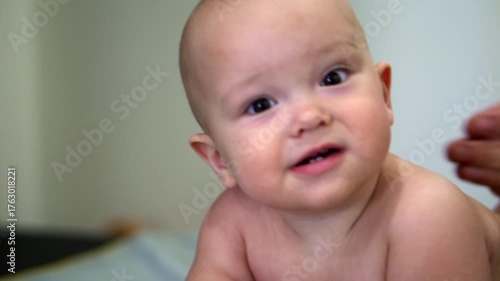 A baby with a bald head is looking at the camera. The baby is wearing a diaper and is laying on a bed