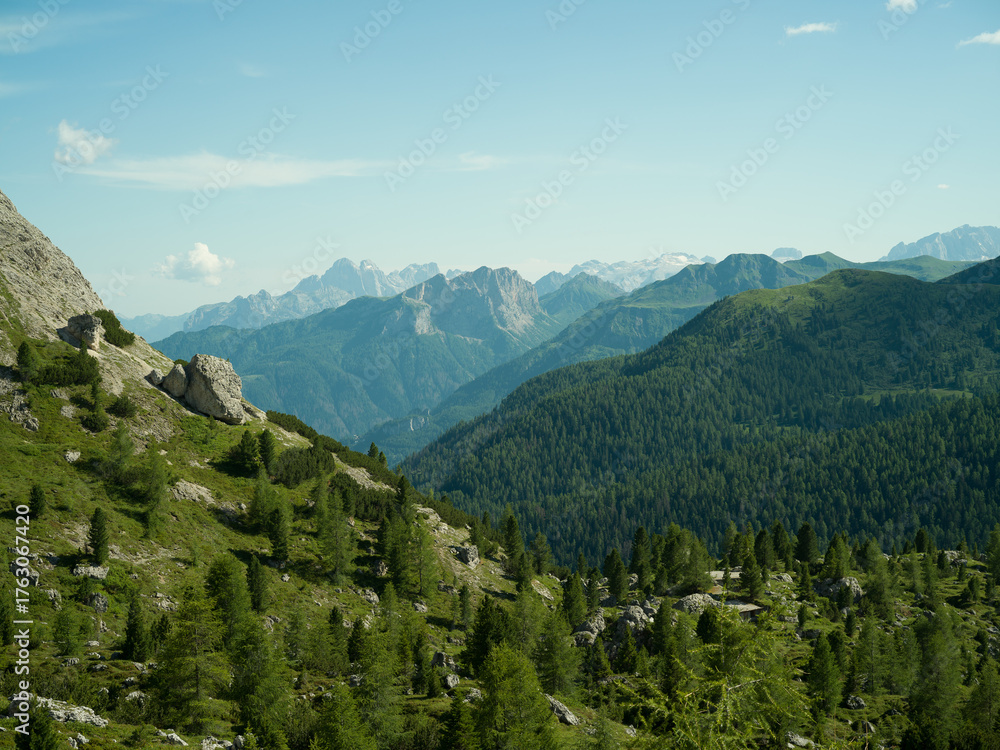 Fototapeta premium Peaceful alpine valley in the Dolomites, Italy. Rolling green hills and pine forests leading to snow-covered peaks in the distance under a soft summer sky — pure mountain serenity.