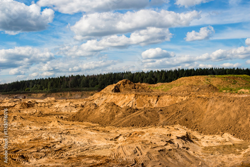Vast open-pit sand quarry under a bright sky, featuring massive earth mounds and distinct tire tracks. It shows an industrial landscape shaped by excavation