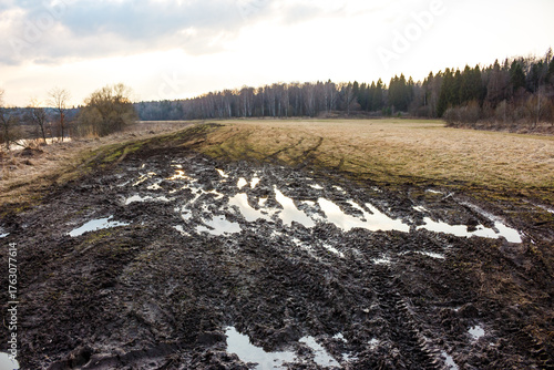 Gnarly, slushy mud track, riddled with puddles and tire ruts, traversing a desolate field towards a distant forest line under an overcast sky