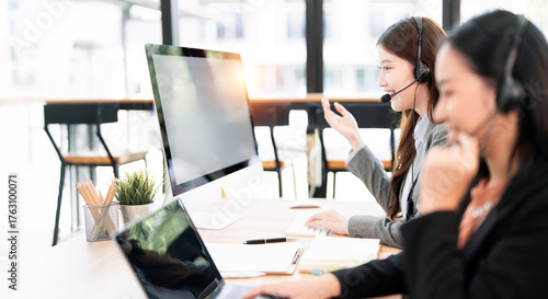 Quadro em tela Two dedicated Asian female call center agents wearing headsets, collaborating and speaking to customers while working on computers in a bright, modern office