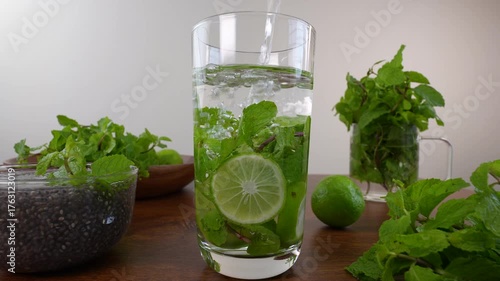 Water pouring into a glass with lemon slices and mint leaves on wooden table.