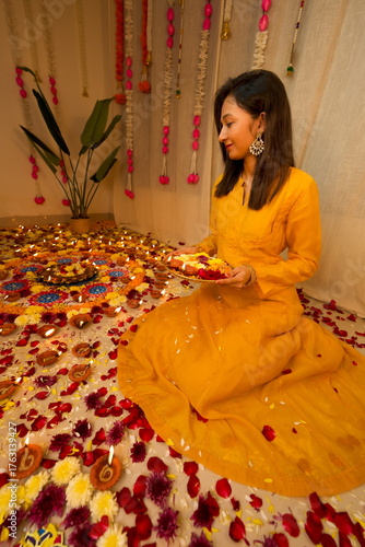 Happy young Indian woman wearing traditional yellow clothes holding plate with oil lamps and flowers celebrating Diwali festival. Festival of lights. Deepawali.