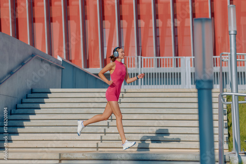 Woman running outdoors on sunny day, wearing headphones, near modern building and steps