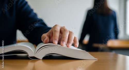 Closeup of a persons hand turning the page of an open book, with another person blurred in the background, suggesting learning or study
