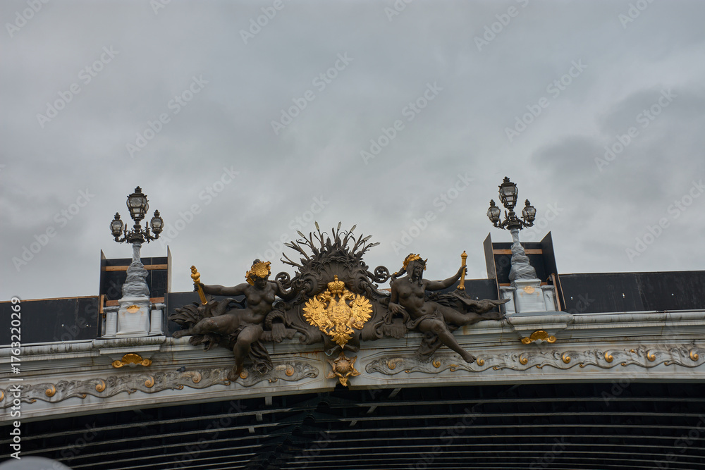 Obraz premium Sculptural detail of the Pont Alexandre III in Paris with golden figures and the imperial emblem against a cloudy sky