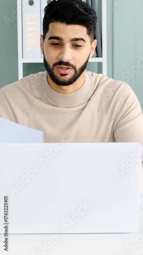 A smiling young adult man speaks and gestures during a video conference call on his laptop and waves goodbye at the end of the meeting.