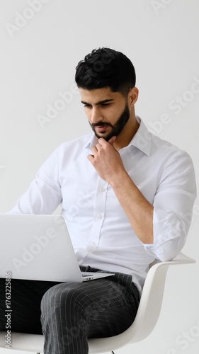 A thoughtful young adult businessman with a beard sits in a modern chair, working and typing on his laptop against a white background.