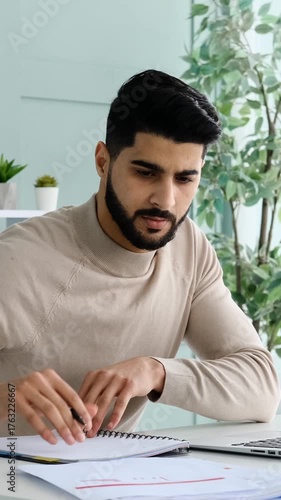 A focused young adult man with a beard works at his desk, analyzing documents, writing notes, and using a laptop in a modern office.