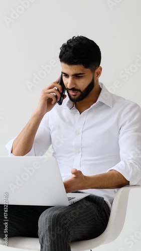 A young adult businessman has a serious conversation on his smartphone, gesturing as he works on his laptop in a modern white studio.