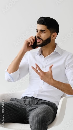 A young adult businessman has a serious conversation on his smartphone, gesturing as he sits in a chair against a white background.