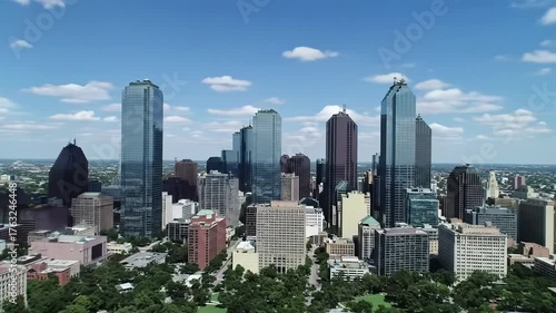Ultra-wide aerial video of Dallas, Texas skyline. Gleaming glass, steel skyscrapers reflect midday sun. Azure sky, white clouds above. Lush city parks, streets below. Thriving metropolis overview