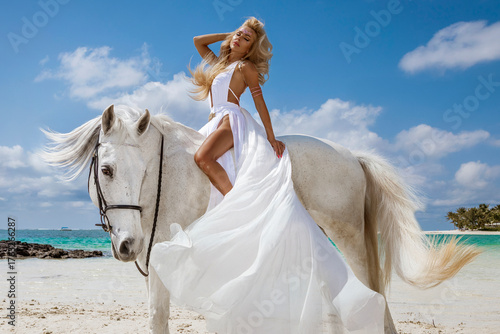 Canvas Print Beautiful woman with long blonde hair wearing a white flowing dress, sitting gracefully on a white horse by the turquoise ocean on a tropical beach in Mauritius