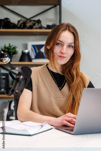 Fotomural A young woman types at a laptop beside a broadcast microphone and notebook in a modern home studio