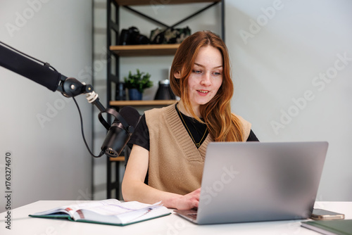 Young woman records at a desk with a laptop and podcast microphone, title in description. She references a notebook and phone as soft daylight and neutral tones set a modern look.