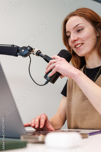 Fotografía Young woman records audio at a tidy desk, speaking into a dynamic mic on a boom arm while working on a laptop in a home studio, daytime lighting and shallow depth of field