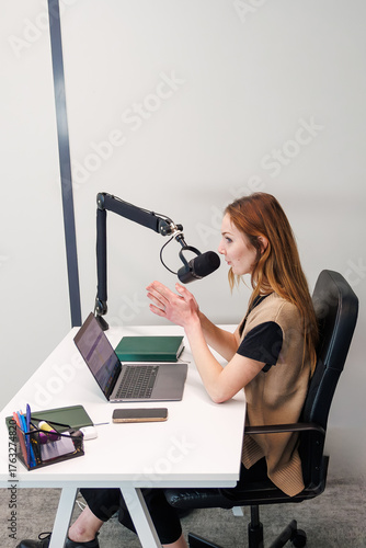 Fotografía A woman with auburn hair speaks into a suspended cardioid mic on a boom arm at a white desk with laptop, tablet, phone, and notebooks in a modern studio during daytime